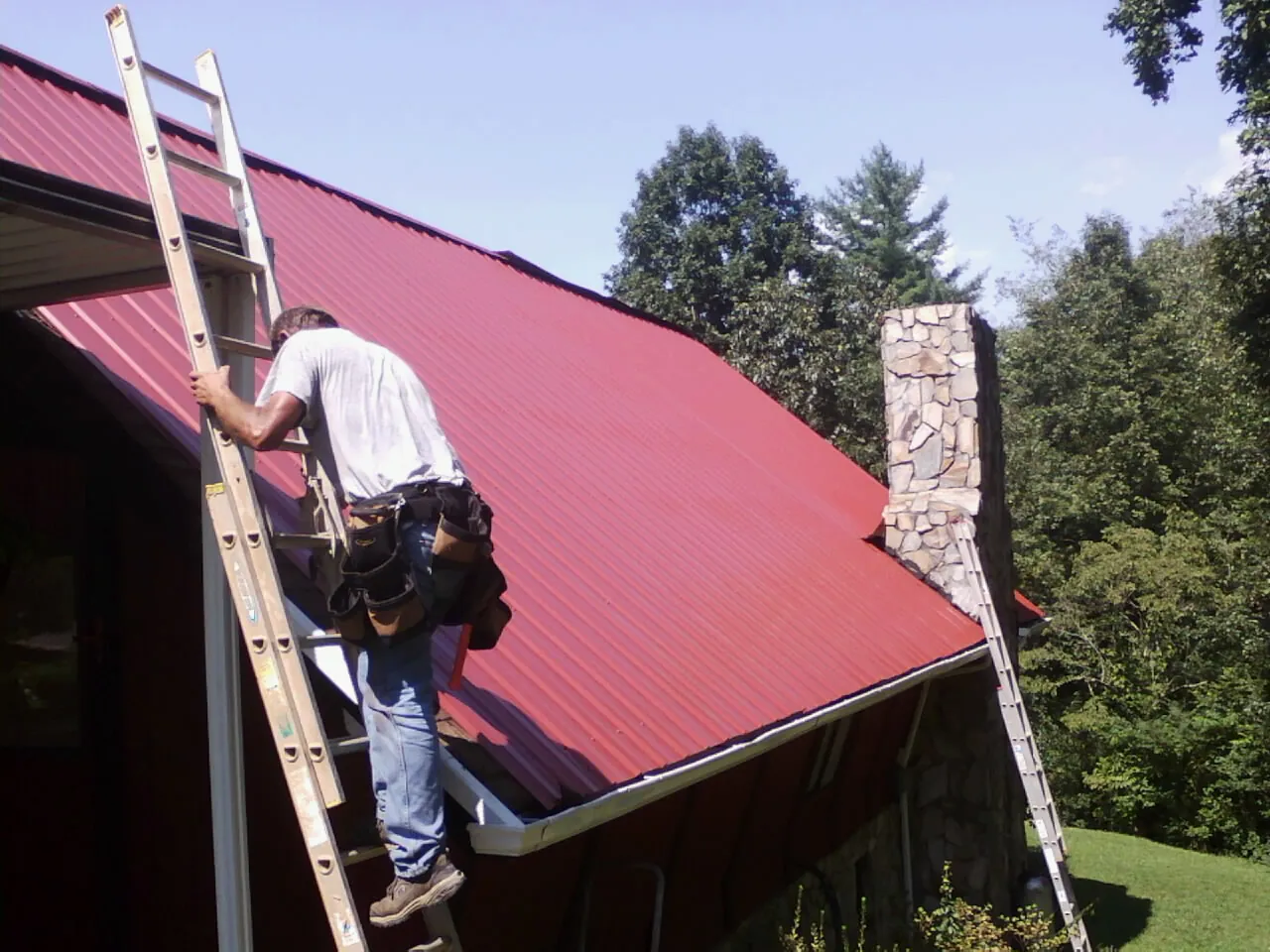 Roofer on a ladder beside a red metal roof and stone chimney — Soffit & Fascia Repair by Roofing Brothers in Spring Hill